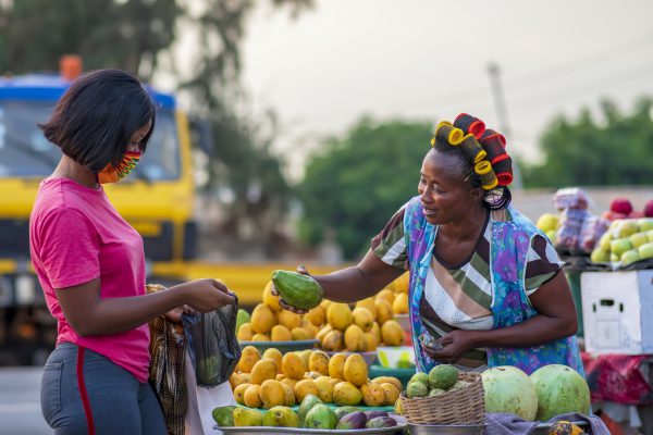 A young African buying fruits from the market and wearing face mask for protection - Receiving a purchased item from a local happy food vendor
